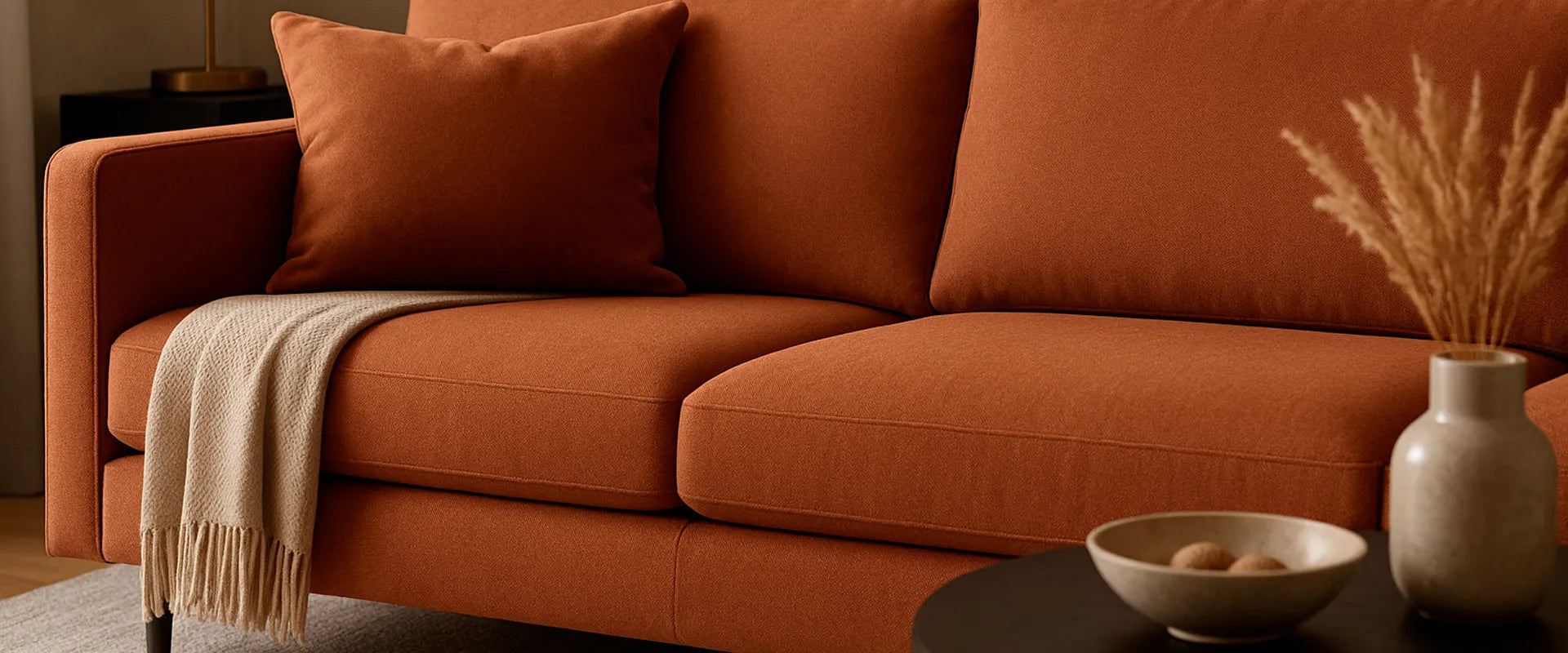Sofá en vista detalle en tela color terracota, con patas en madera tono oscuro, cojín terracota y manta beige, elementos decorativos como mesa auxiliar y jarrón, ambiente sofisticado y minimalista - All Star Colombia Muebles